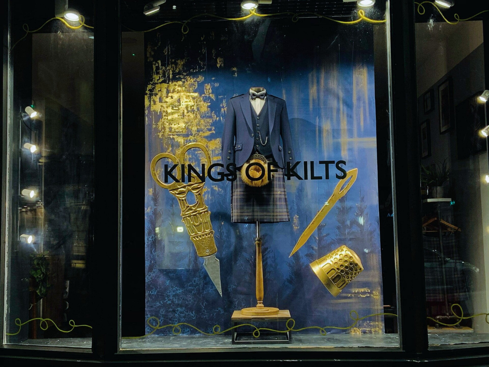 Storefront of a kilt store, with a forest background and faux giant gold scissors, needle and thimble